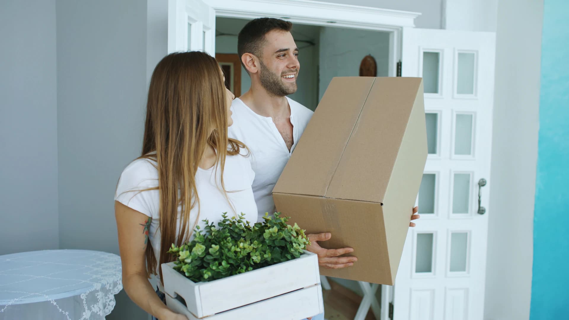 Couple carrying moving boxes and a plant into their new home